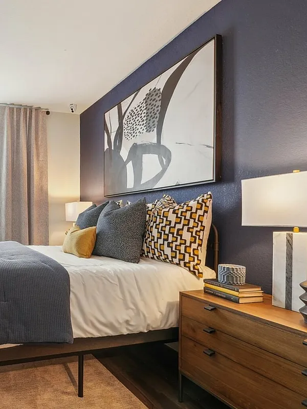 Modern bedroom with stylish decor, featuring a navy accent wall, elegant artwork, and a cozy double bed with vibrant cushions and wooden nightstands.