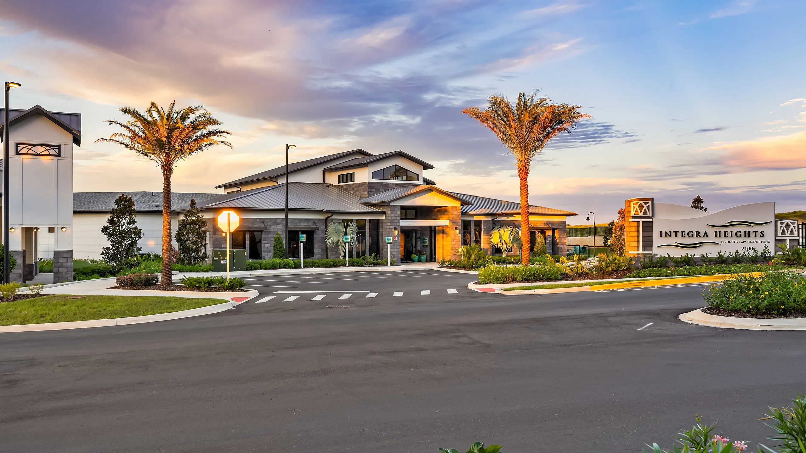Elegant entrance of Integra Heights, showcasing modern architecture surrounded by lush landscaping and palm trees, under a vibrant sunset sky.
