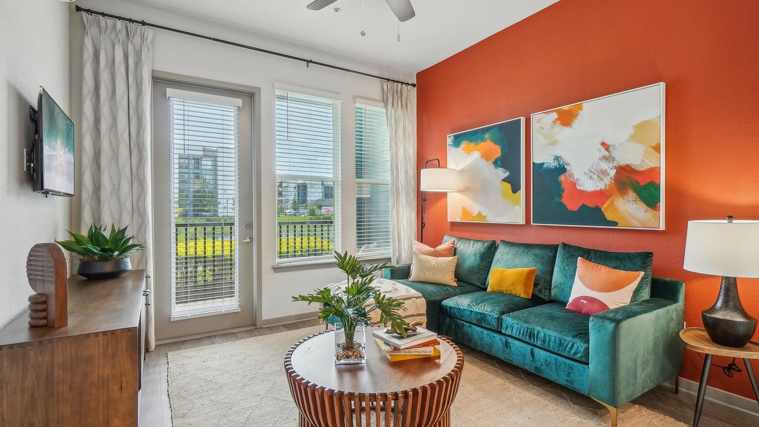 Vibrant living room with bold orange accent wall, modern teal sofa, abstract artwork, and expansive windows offering abundant natural light.