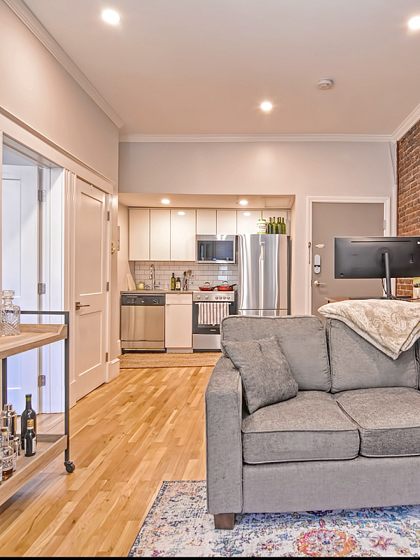 Chic apartment living room with a cozy gray sectional sofa, exposed brick wall, and modern kitchen, featuring hardwood floors and stylish lighting.