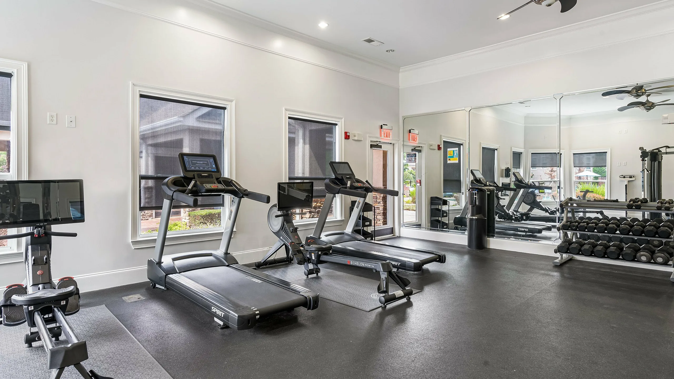 Modern gym area with state-of-the-art equipment, including treadmills, a rowing machine, and a full set of free weights, enhanced by natural light and large mirrors.