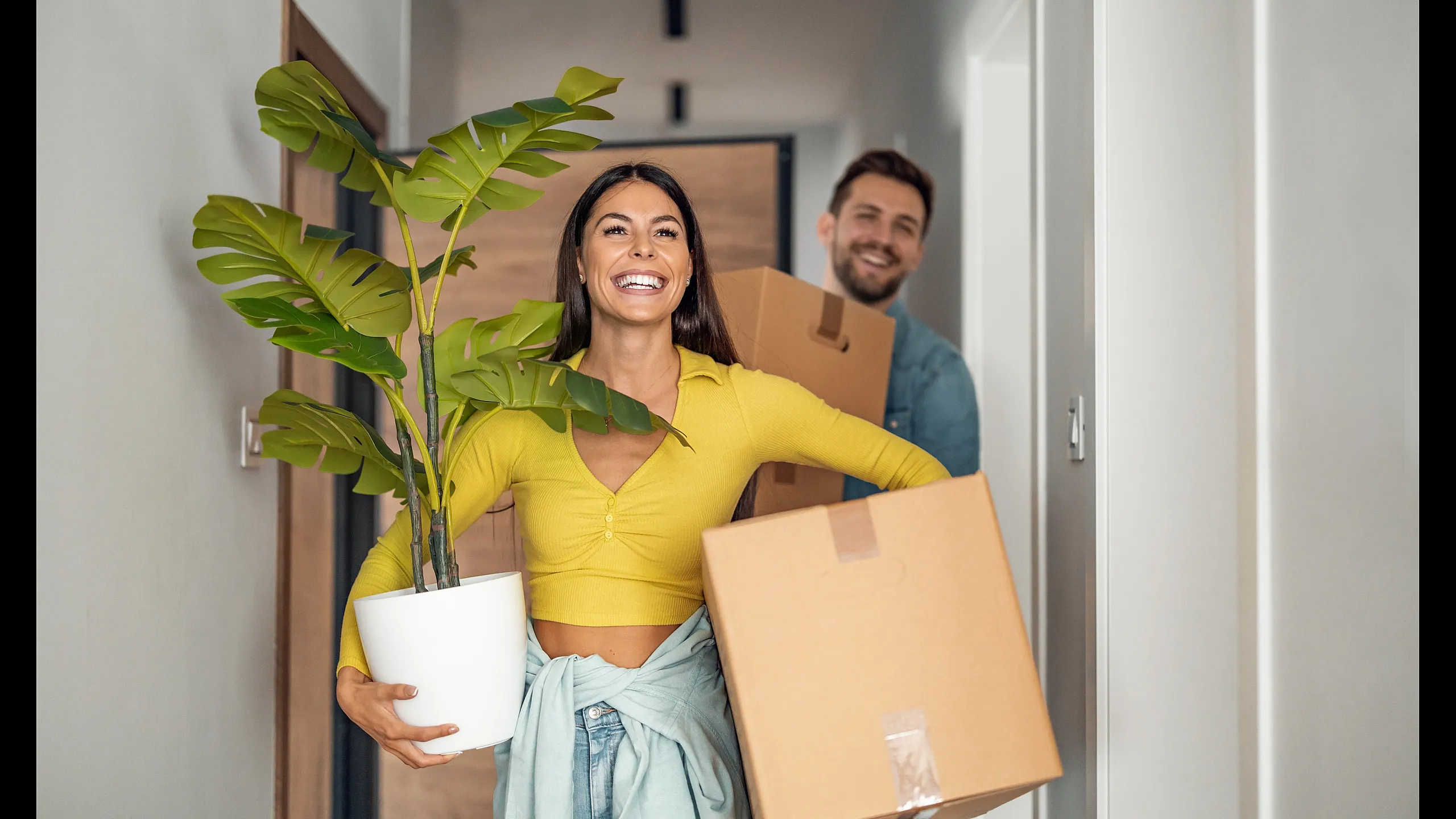 Joyful couple moving into a new home, carrying a potted plant and boxes, symbolizing fresh beginnings and excitement.