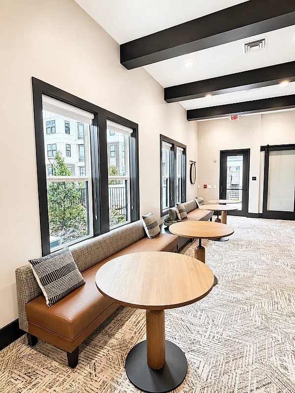 Modern apartment lounge with stylish seating, round wooden tables, and large windows offering abundant natural light.
