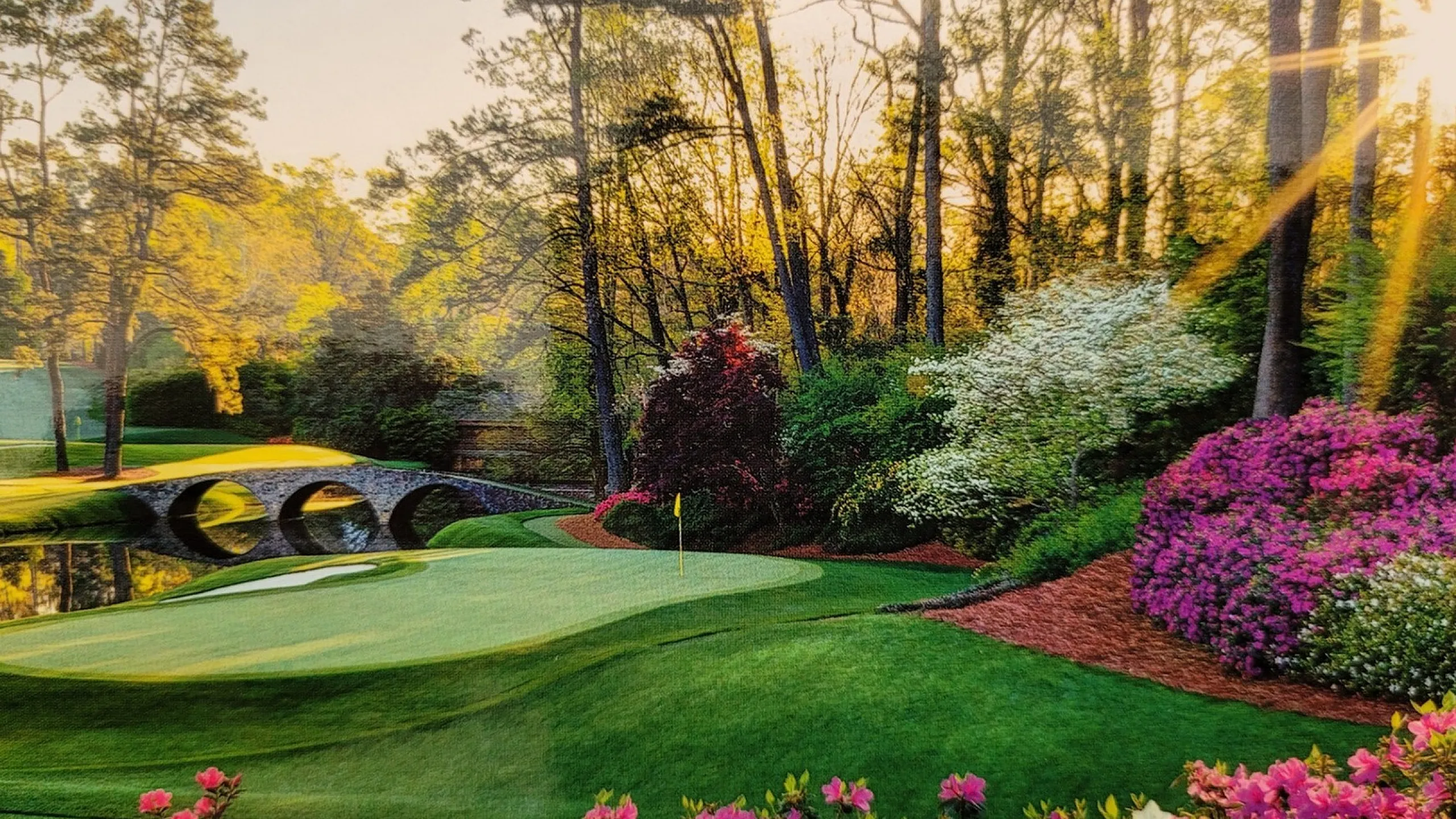 Picturesque landscape with vibrant flowers, a serene pond, and a stone bridge bathed in golden sunlight.