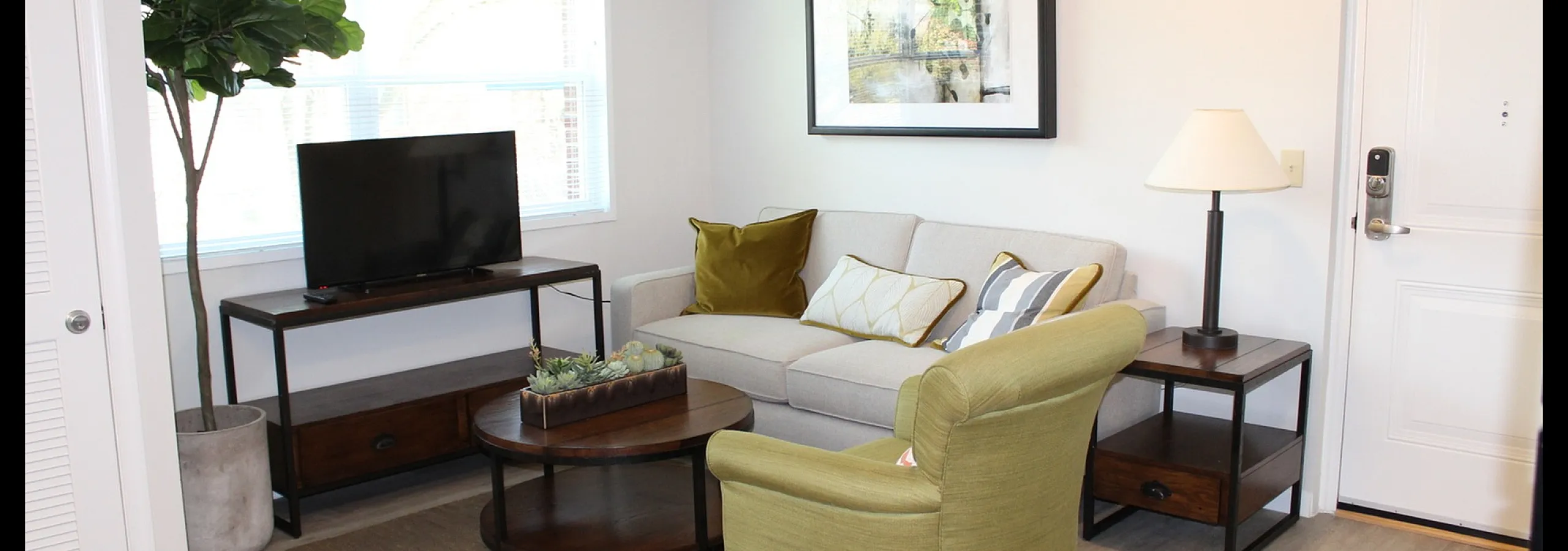 Inviting living room with modern decor featuring a cozy beige sofa, stylish green armchair, and a sleek TV stand by a large window, complemented by a chic floor plant and elegant artwork.