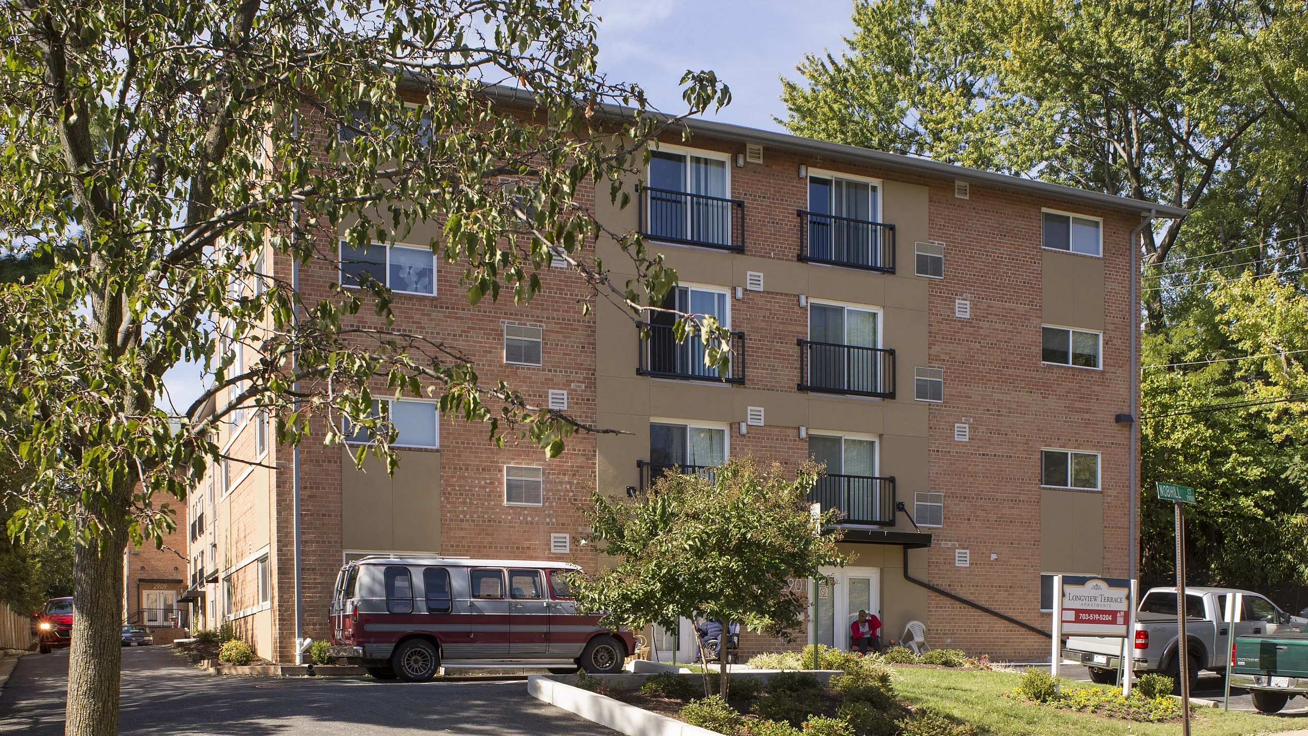 Charming brick apartment building surrounded by lush greenery, offering bright balconies with modern railings and ample parking space.