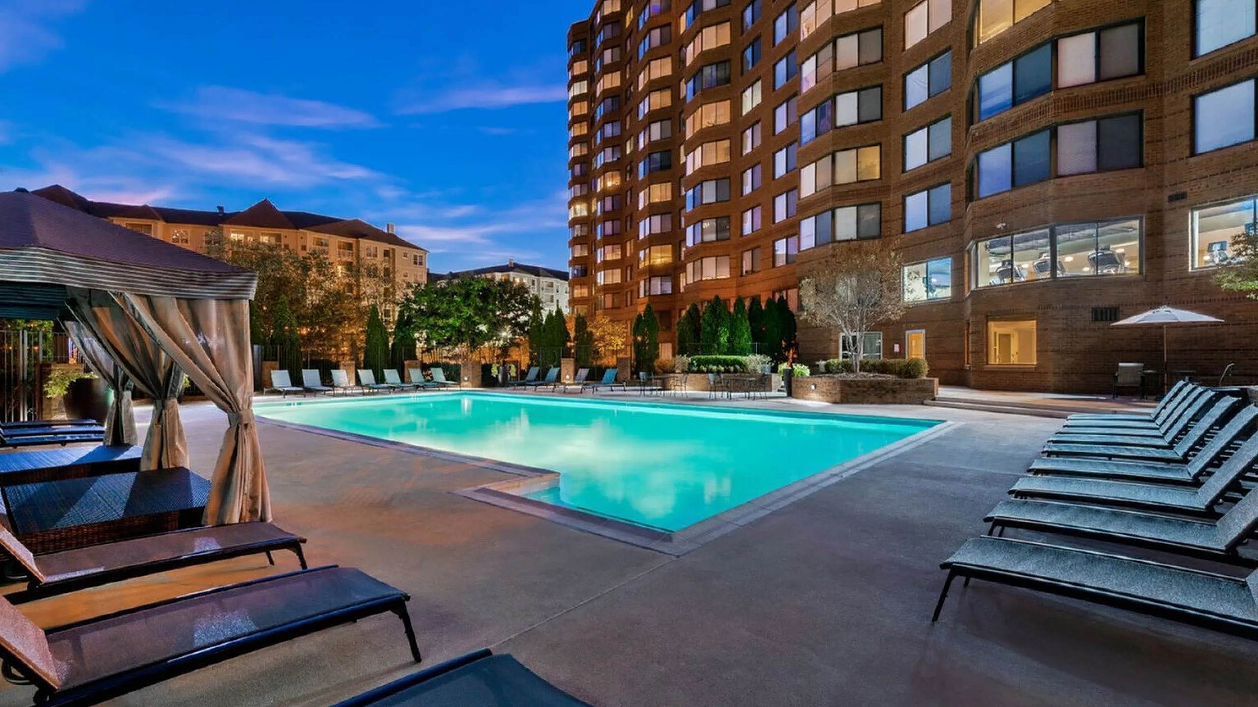 Luxurious apartment complex featuring a beautifully illuminated outdoor pool area with elegant lounge chairs and scenic views at dusk.