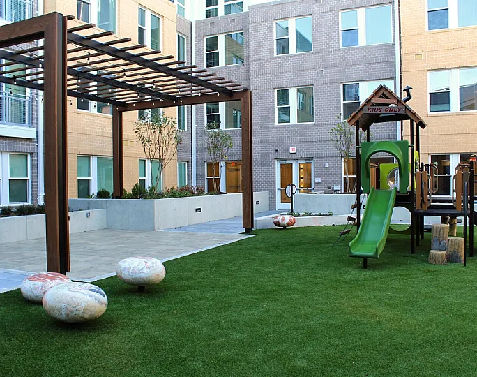 Modern urban courtyard featuring a vibrant playground with green slide, wooden pergola, and artistic stone seating, perfect for family enjoyment.