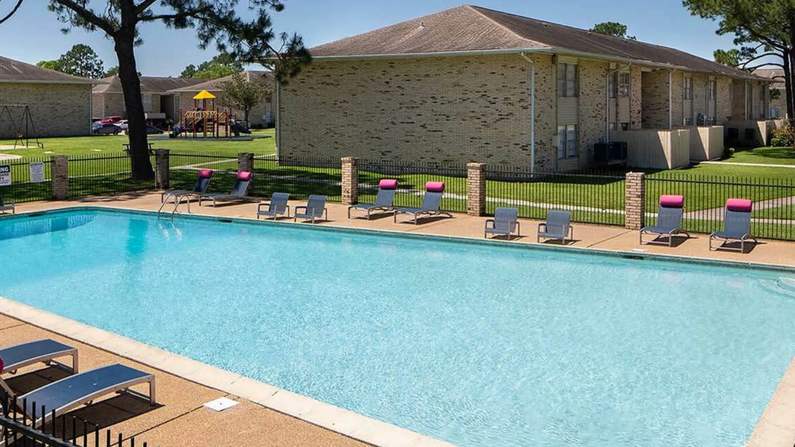 Inviting apartment pool with surrounding lounge chairs and lush greenery on a sunny day.