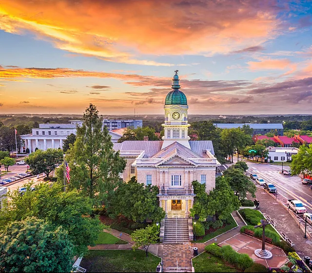 Aerial view of a historic building with a clock tower surrounded by lush greenery under a vibrant sunset sky.