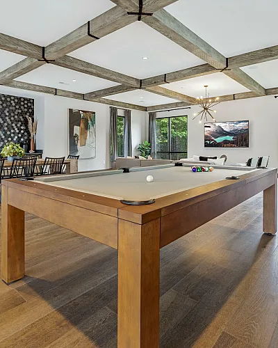 Luxurious open-concept living area featuring a stylish pool table, elegant dining space, and expansive windows with lush greenery views.
