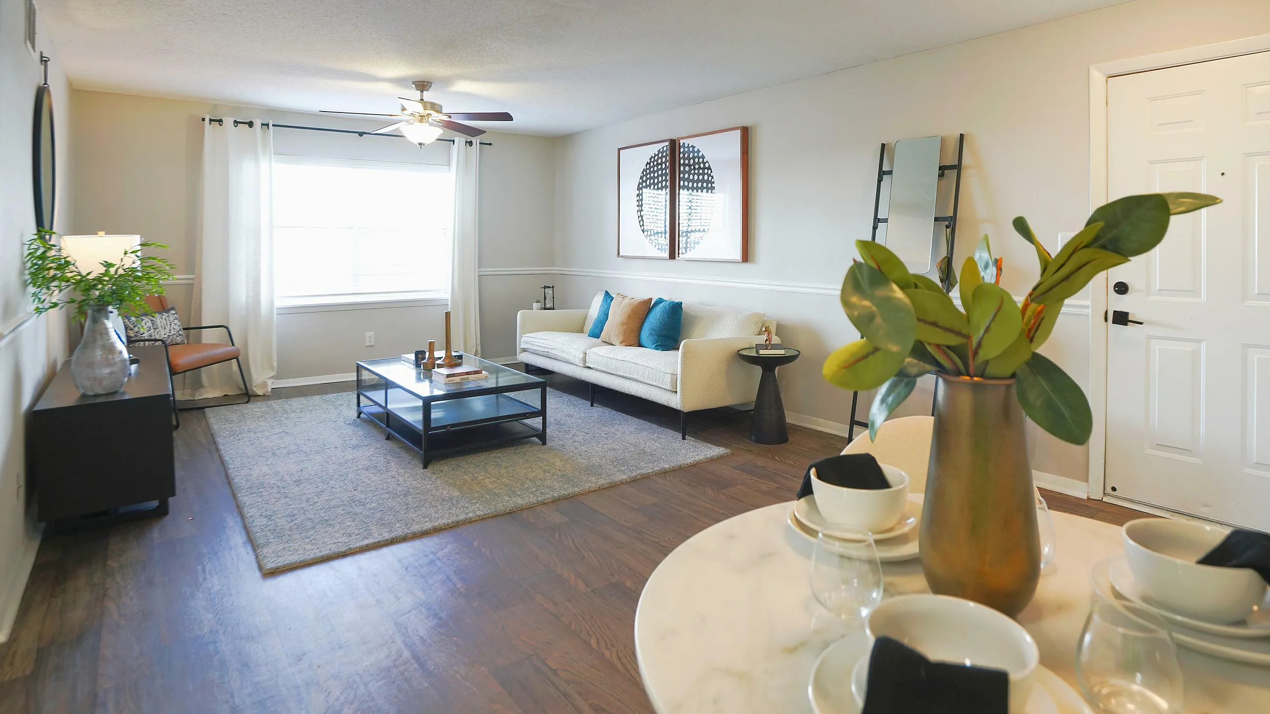Inviting living room with warm wooden floors, a cozy white sofa with vibrant cushions, a modern glass coffee table, and elegant decor accents under natural light.
