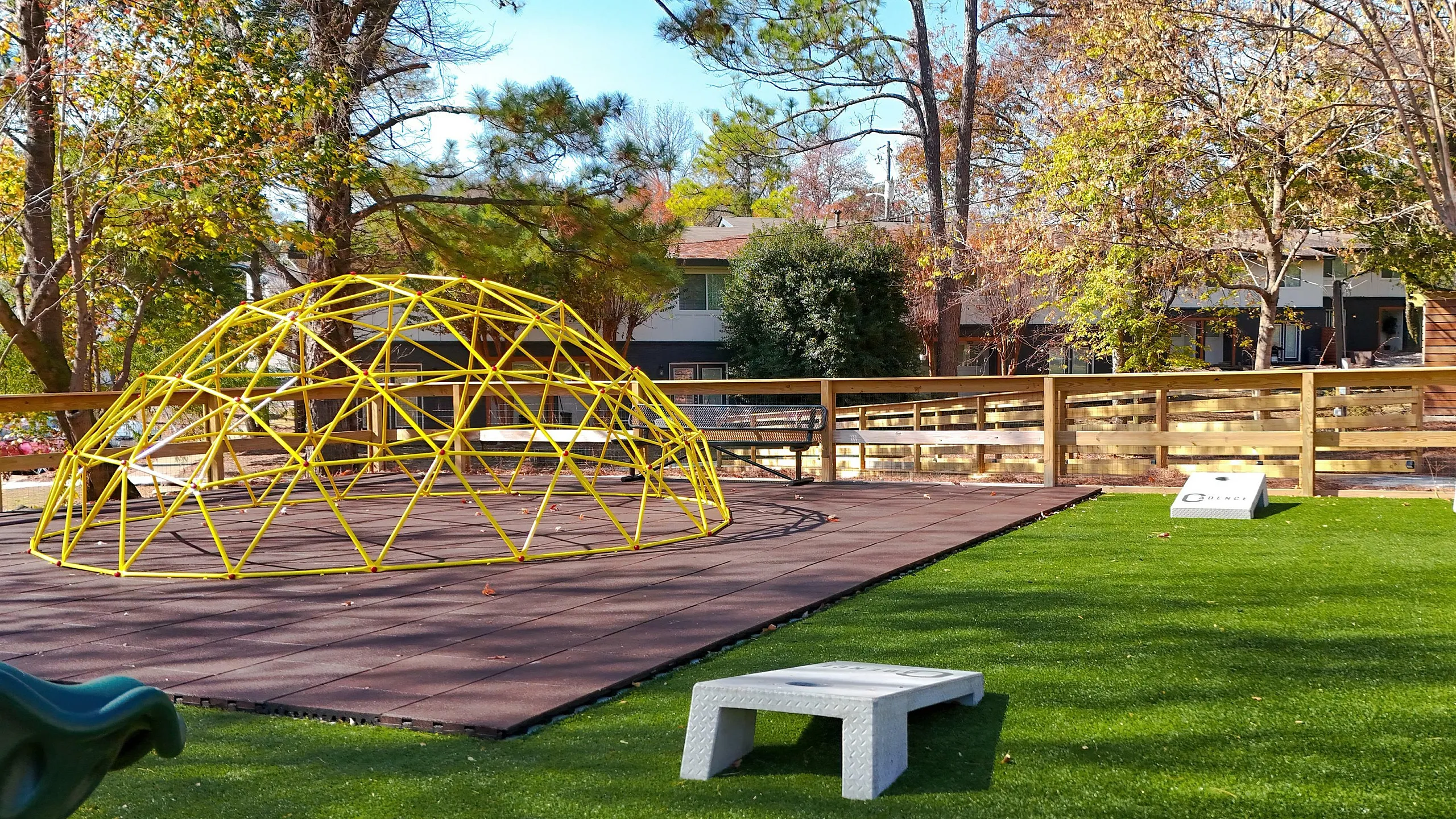 Charming outdoor play area with vibrant green artificial turf, featuring a unique yellow geodesic climbing dome, surrounded by mature trees and a wooden fence.