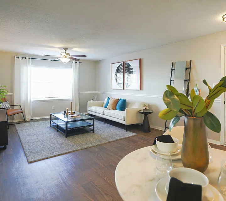 Bright and inviting living room featuring a cozy white sofa with colorful cushions, elegant coffee table, and large window with abundant natural light, complemented by a stylish dining area with a modern table setting and decorative plant.