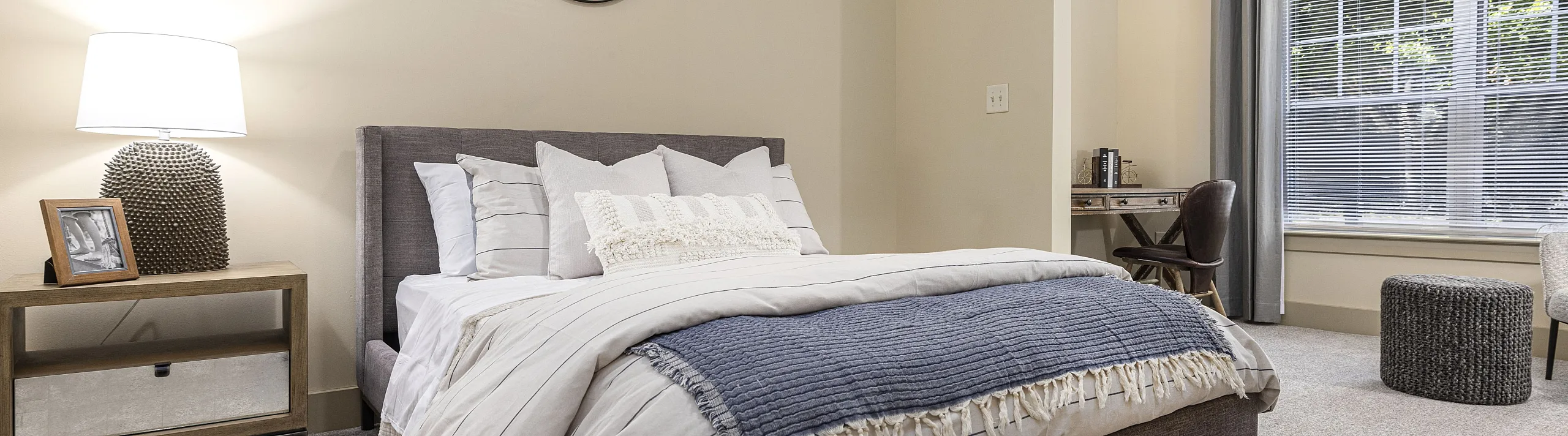 Cozy bedroom with plush carpeting, featuring a stylish bed, elegant side table with lamp, and sunny window with a serene view.