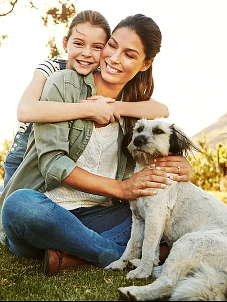 Joyful mother and daughter embracing their pet dog outdoors on a sunny day, surrounded by lush greenery.
