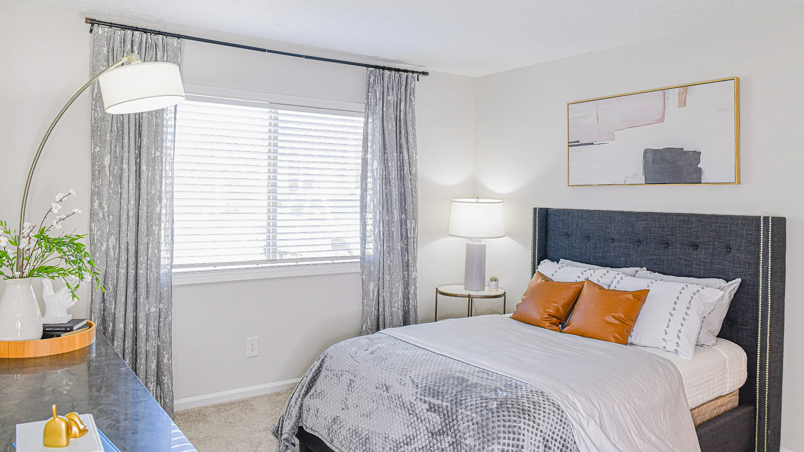 Bright and inviting bedroom with contemporary decor, featuring a plush bed with chic linens, elegant floor lamp, abstract art, and ample natural light from a large window.