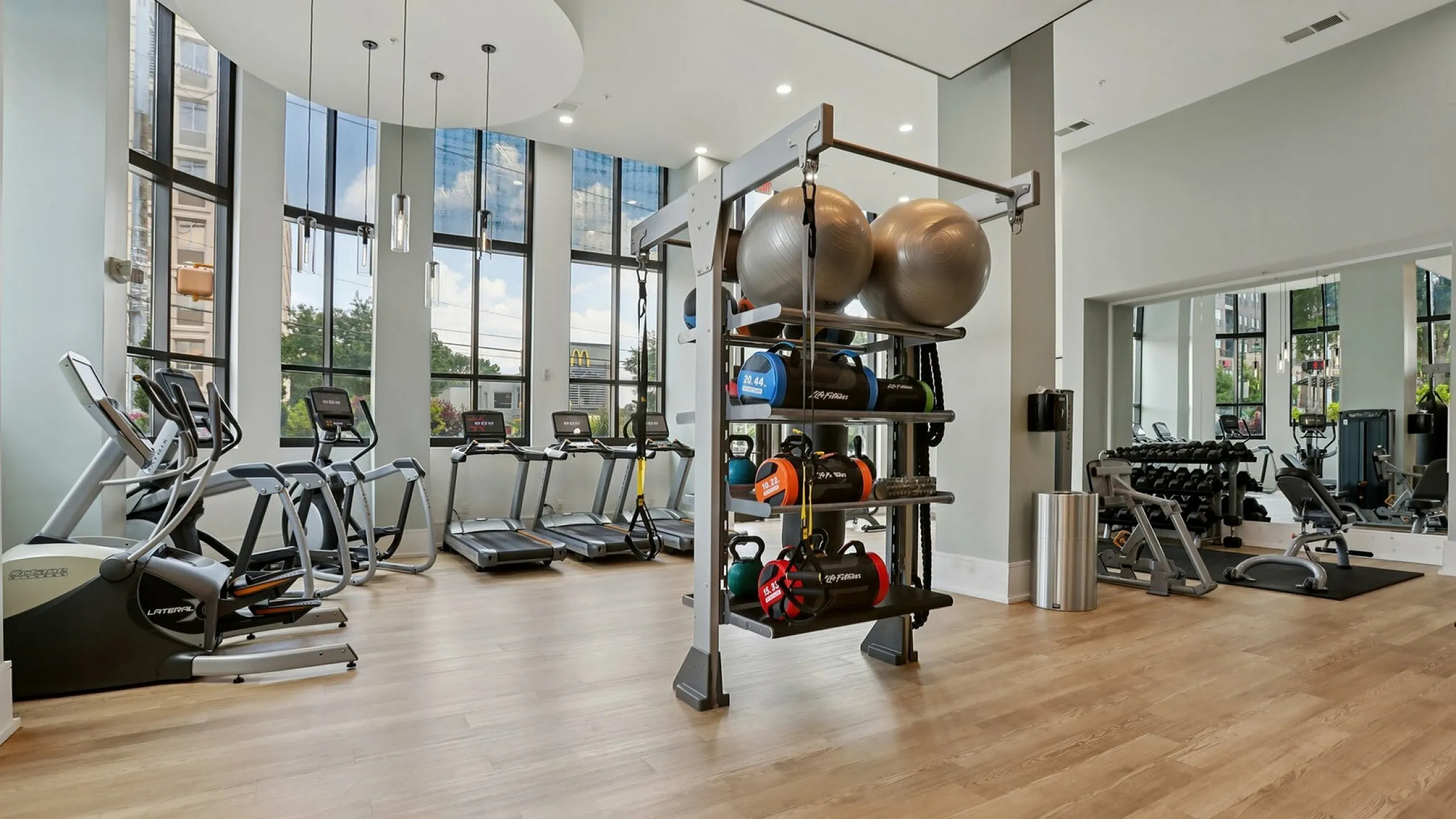 Modern fitness center with large windows, featuring treadmills, weight racks, and a variety of gym equipment in a spacious and well-lit environment.