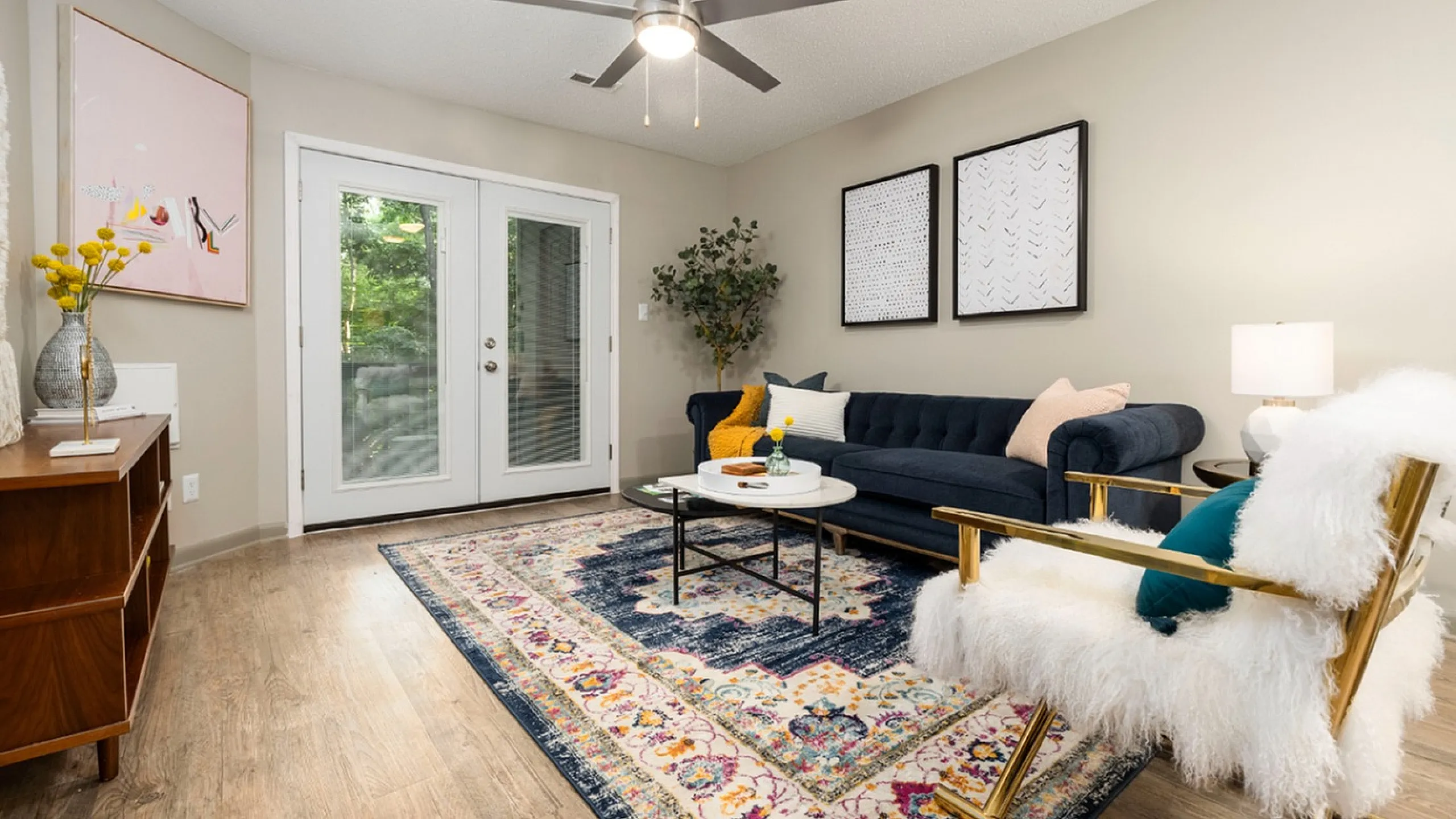 Chic living room with elegant decor, plush navy sofa, fluffy white armchair, and vibrant area rug on hardwood floor, framed by stylish art and a light-filled glass door.
