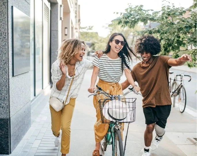 Vibrant urban scene with three friends, two walking and one riding a bicycle, enjoying a sunny day on a city sidewalk.