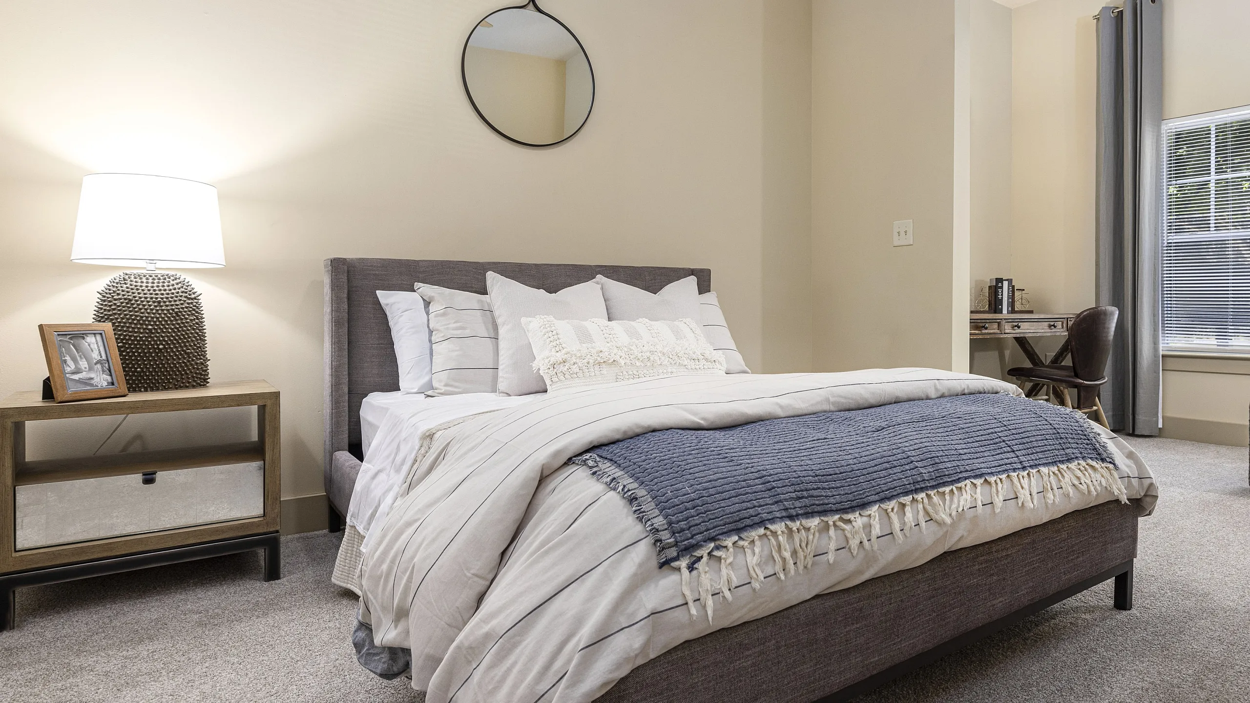 Cozy bedroom with plush carpeting, elegant decor, and ample natural light from a large window, showcasing contemporary furnishings and a stylish workspace.