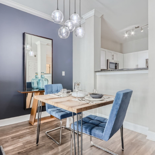 Contemporary dining area with sleek blue chairs, elegant hanging lights, and a chic mirrored accent, adjacent to a modern white kitchen.