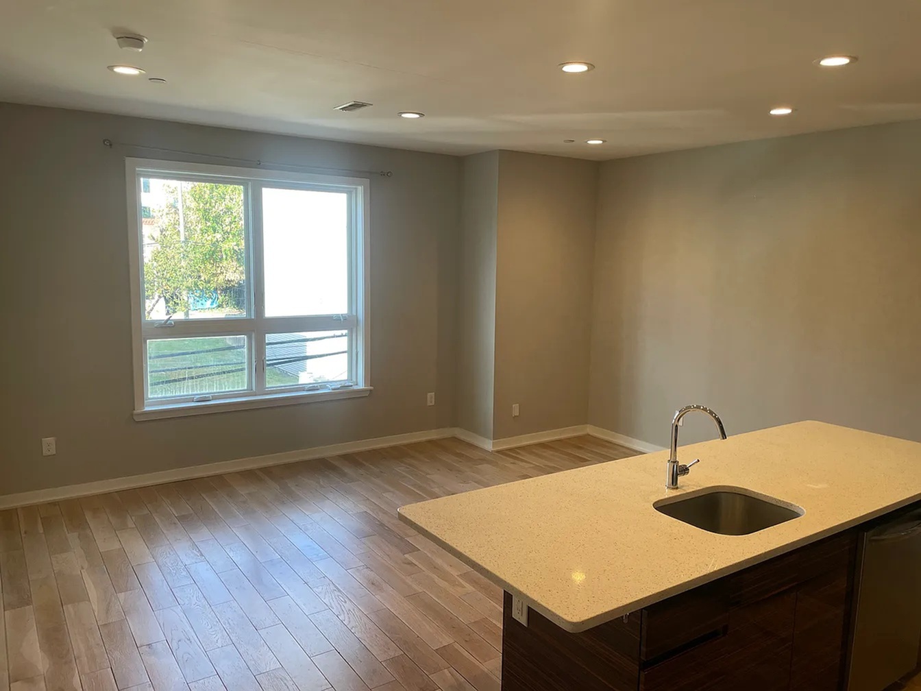 Bright and spacious living area with modern hardwood flooring, large window offering natural light, and a sleek kitchen island with sink.