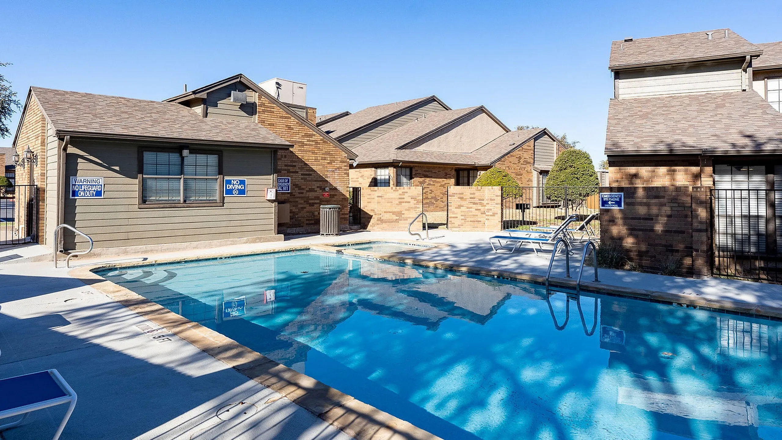 Charming apartment complex with a refreshing outdoor pool, inviting sun loungers, and modern brick architecture under a clear blue sky.