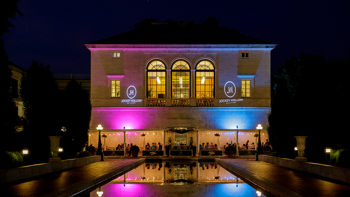 Elegant illuminated building at night showcasing vibrant pink and blue lighting, with a lively dining area reflected in a tranquil water feature.