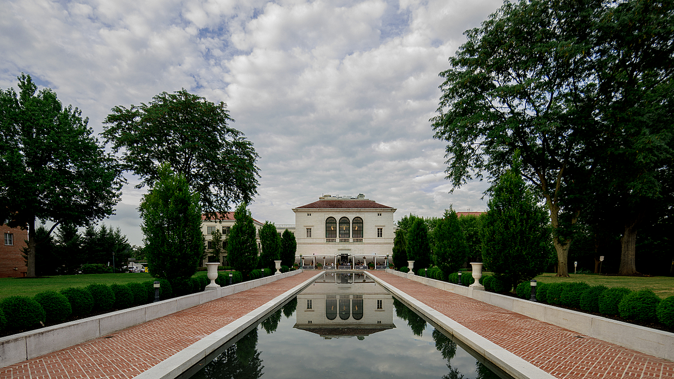Elegant estate with a grand entrance reflected in a tranquil water feature, surrounded by lush greenery and classic architecture.
