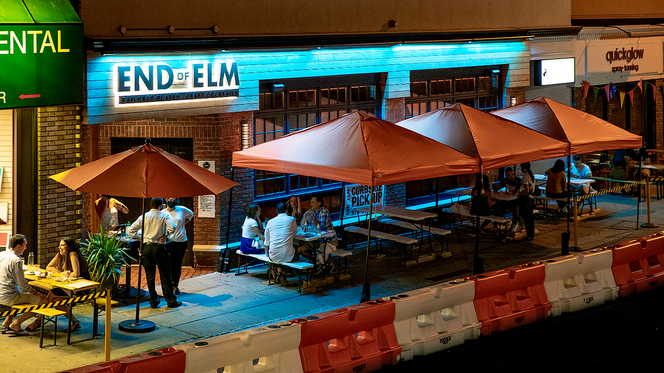 Vibrant night scene at End of Elm with outdoor seating under warm orange umbrellas, lively conversations, and a welcoming ambiance.