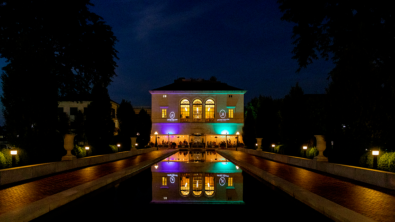 Elegant villa illuminated at night with vibrant lights, reflecting in a serene water feature, surrounded by lush greenery.