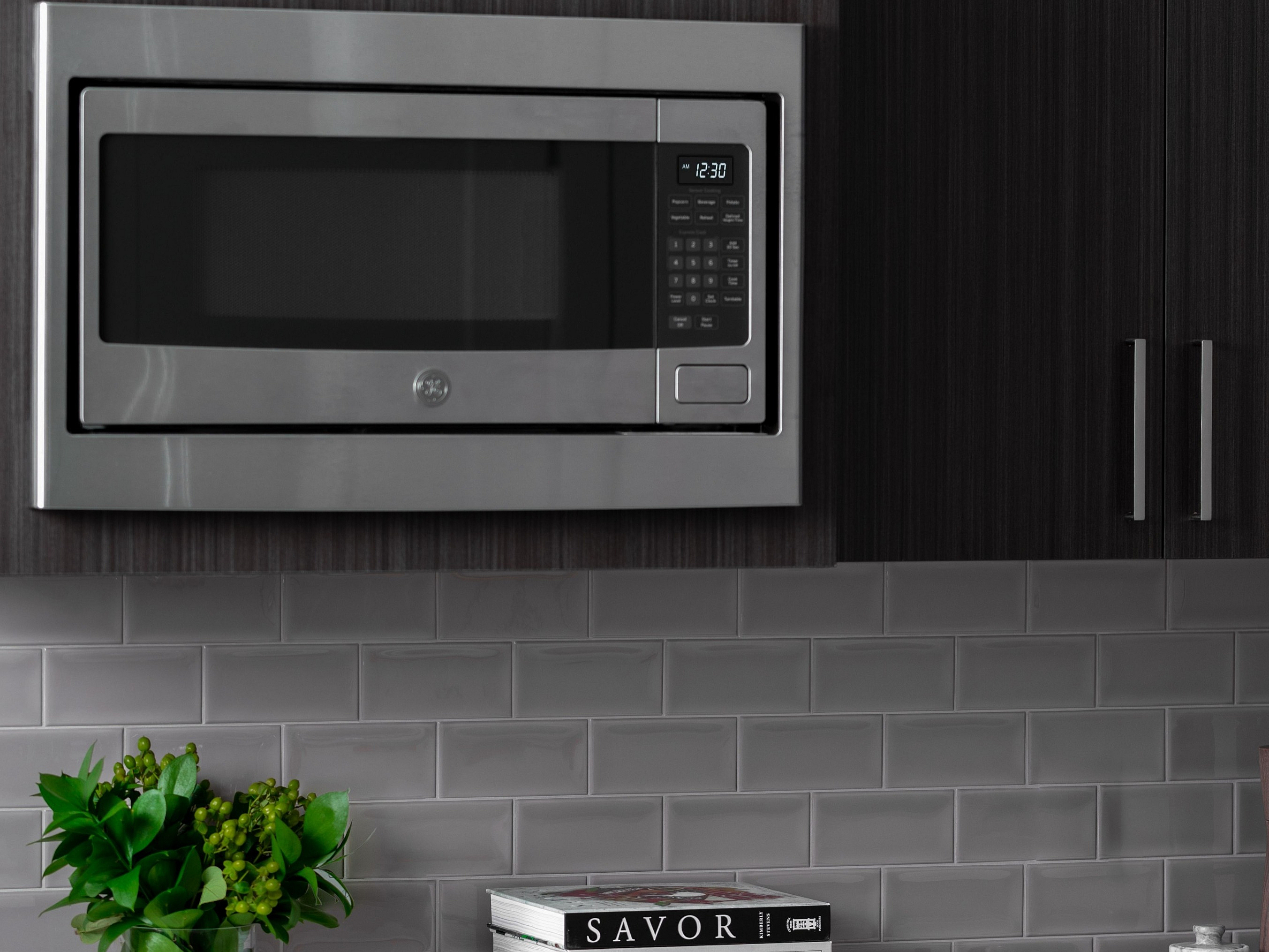 Elegant kitchen featuring sleek stainless steel microwave, modern dark wood cabinetry, and stylish subway tile backsplash, accented with decorative plants and cookbooks.