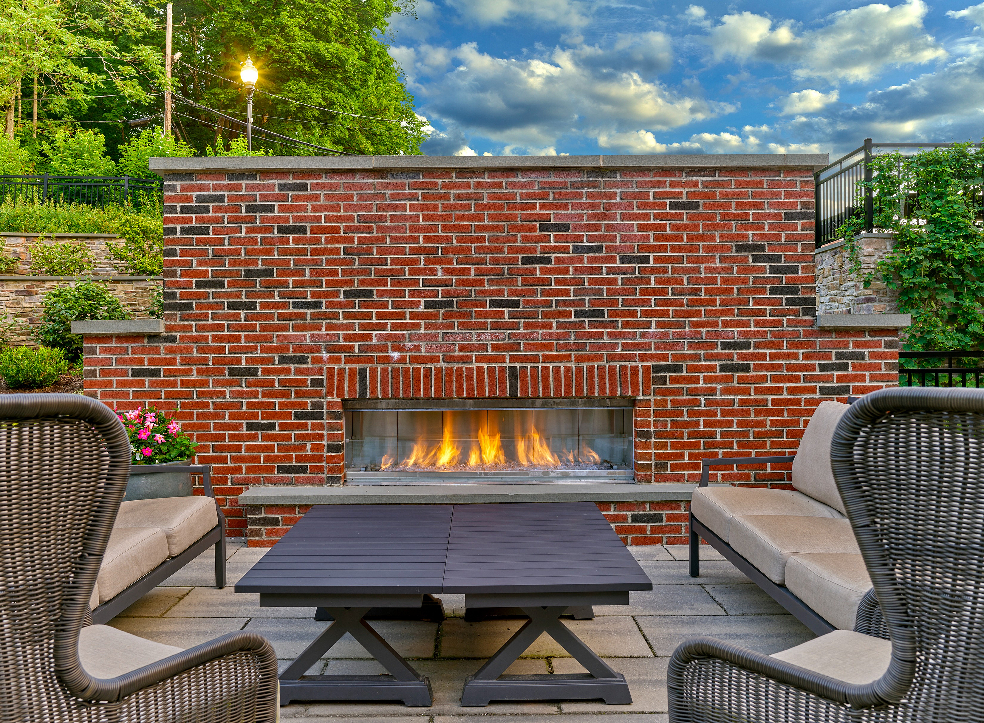 Chic outdoor patio with cozy seating, modern brick fireplace, and lush green surroundings under a serene sky.