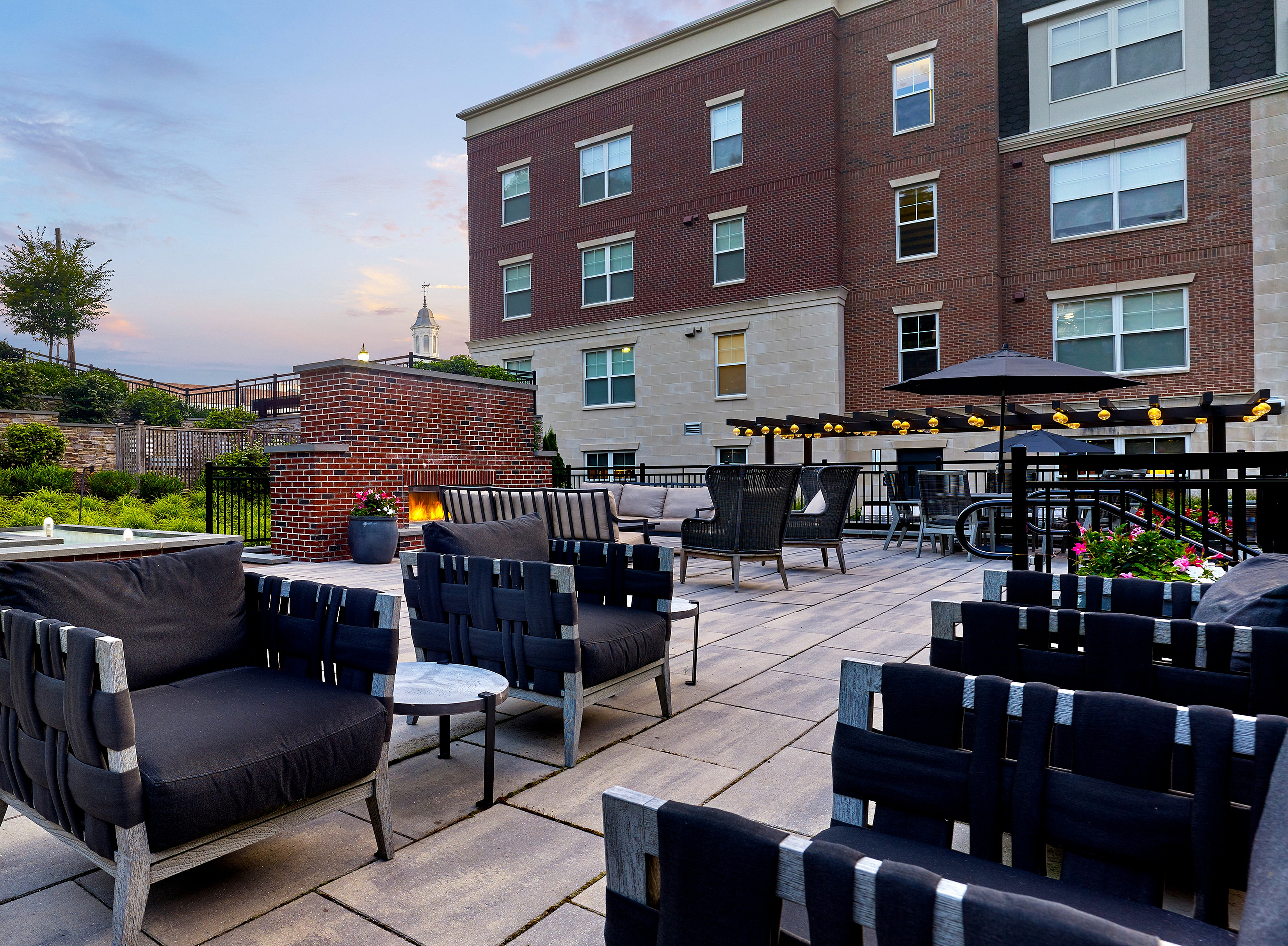 Elegant outdoor patio space with modern seating, cozy fire pit, and ambient lighting against a charming brick building backdrop.