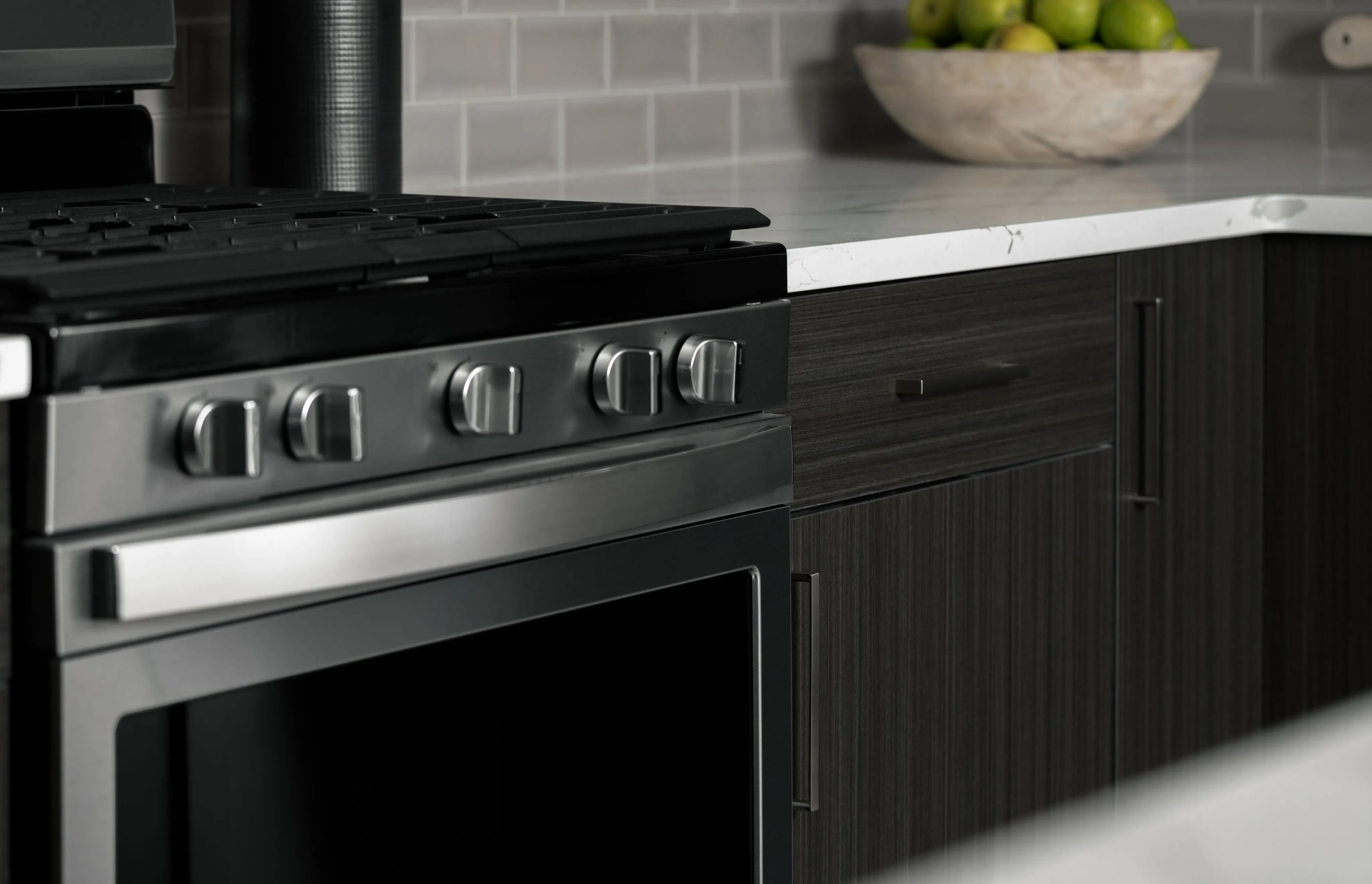Modern kitchen showcasing a sleek stainless steel oven, dark wood cabinets, and stylish backsplash, with a bowl of fresh green apples on the marble countertop.
