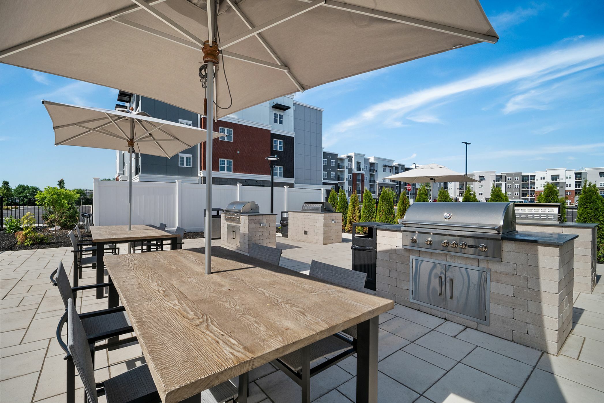 Inviting outdoor patio with elegant wooden tables and umbrellas, featuring modern stainless steel grills, surrounded by lush greenery and contemporary apartment buildings.