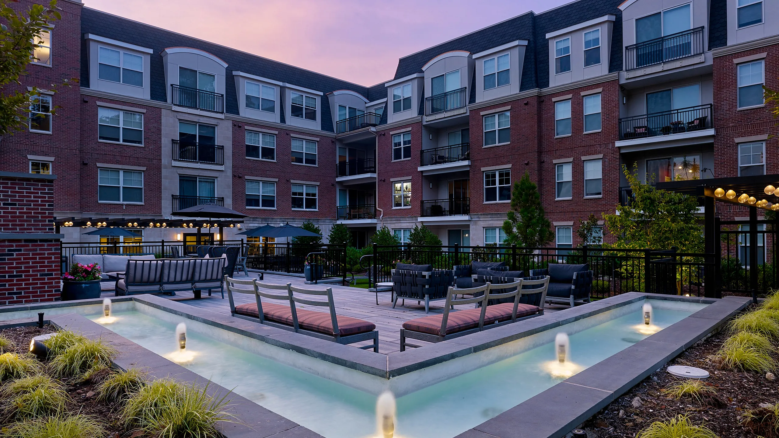 Elegant apartment courtyard featuring modern seating, ambient lighting, and a serene water feature at dusk.