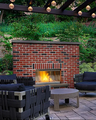 Chic outdoor seating area with cozy woven chairs around a glowing brick fireplace, under a stylish pergola adorned with soft lighting, surrounded by lush greenery.