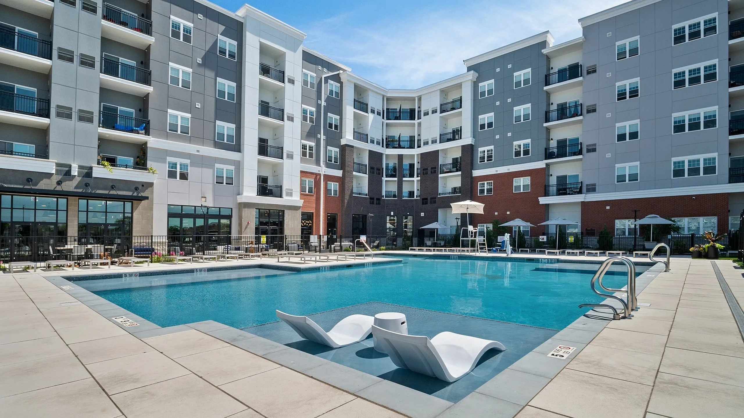 Modern apartment complex with a luxurious outdoor pool and stylish poolside seating, surrounded by multi-story buildings and sunny blue skies.