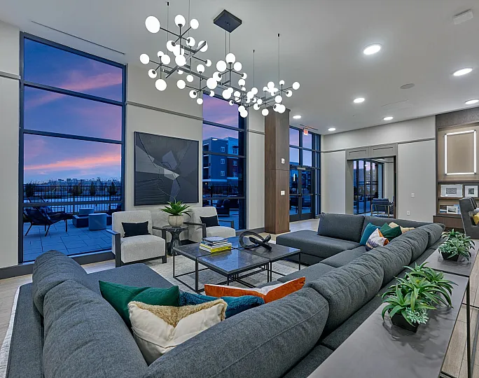 Modern living space with stylish gray sofas, vibrant cushions, artistic chandelier, and panoramic floor-to-ceiling windows showcasing a stunning sunset view.