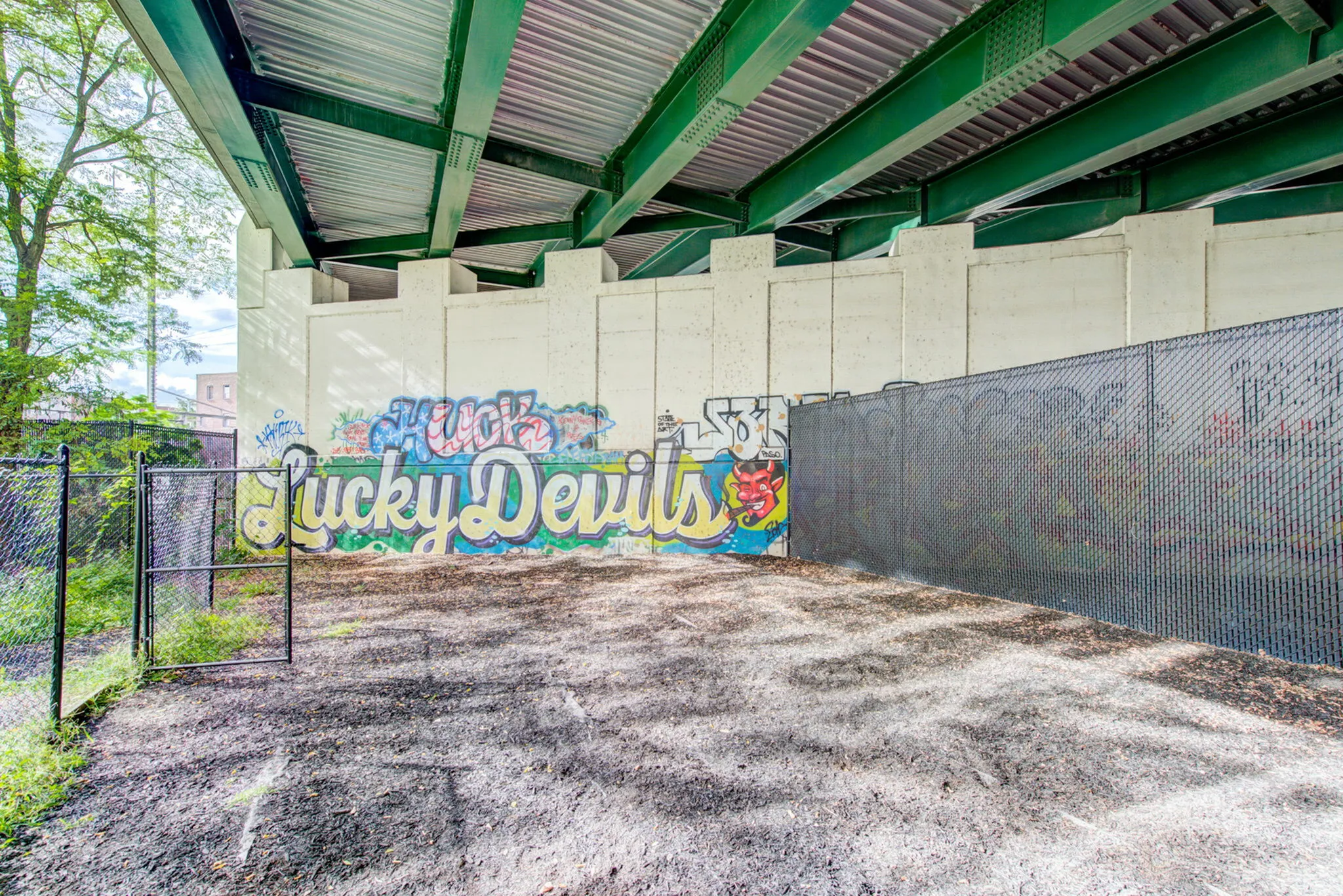 Vibrant urban scene with colorful \'Lucky Devils\' graffiti under a modern bridge, surrounded by lush greenery and chain-link fencing.