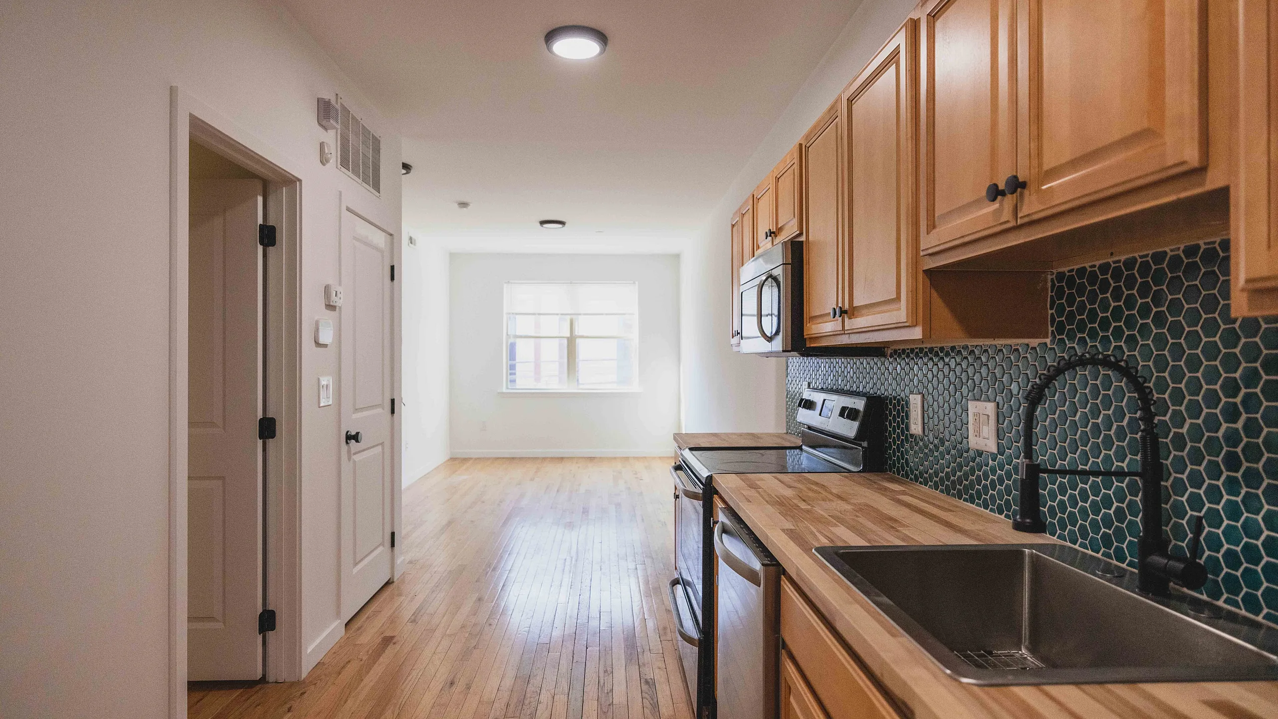 Bright kitchen space with sleek wooden cabinets, modern appliances, and stylish hexagonal backsplash leading into a sunlit living area.