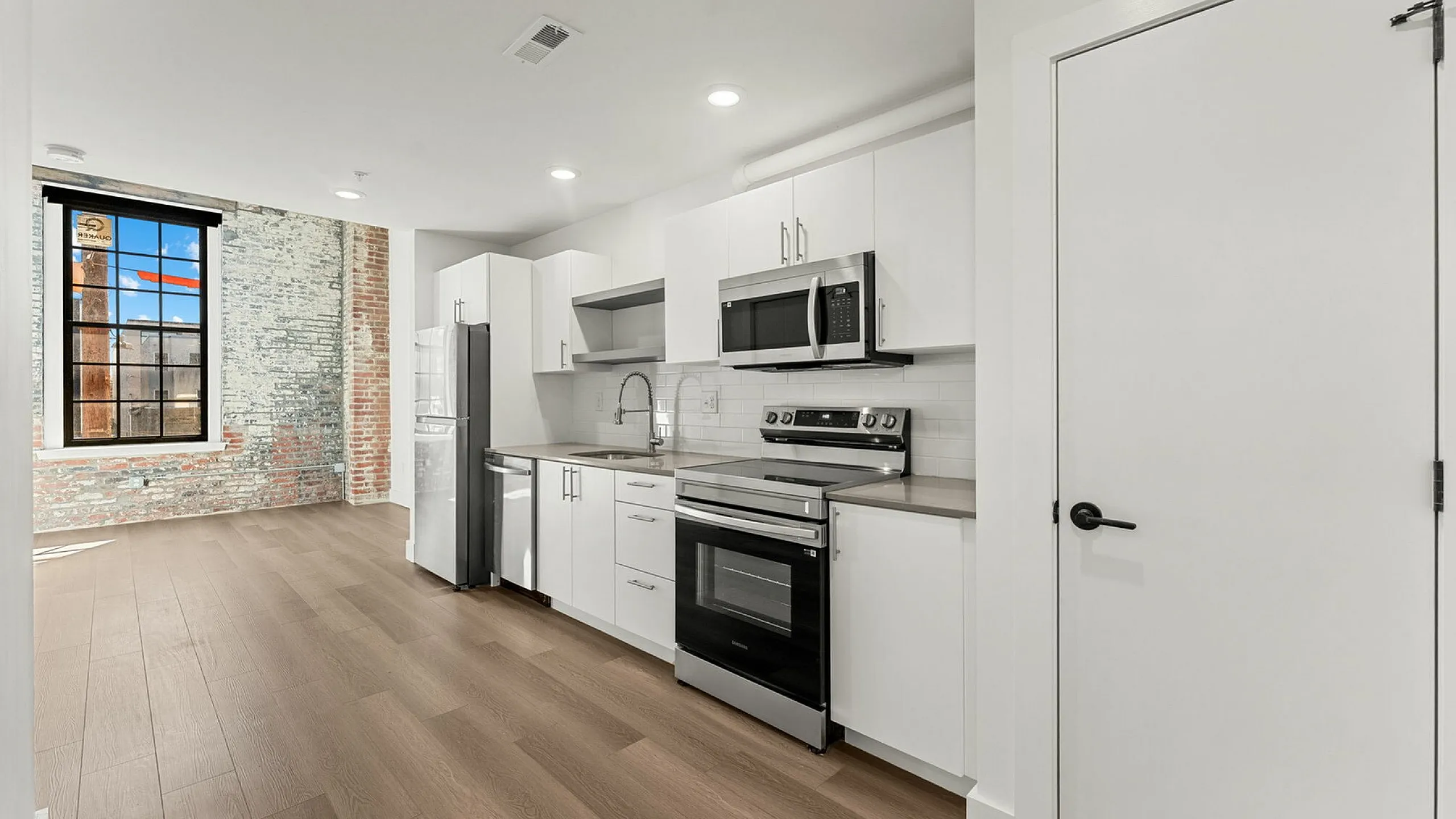 Modern kitchen with sleek white cabinetry, stainless steel appliances, and a stylish open layout, featuring an exposed brick accent wall and large window for natural light.