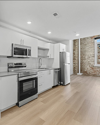 Modern kitchen with sleek stainless steel appliances, white cabinetry, and warm wooden flooring, complemented by an exposed brick accent wall and natural light.