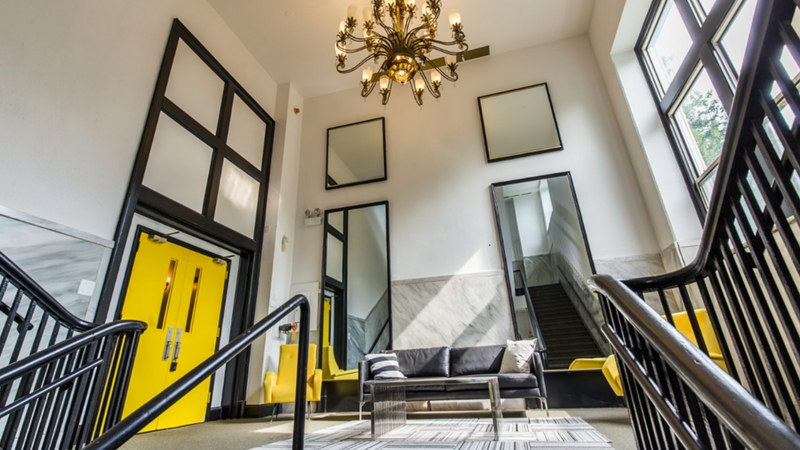 Spacious and elegant apartment lobby with a grand chandelier, vibrant yellow doors, cozy seating, and large mirrors reflecting natural light.