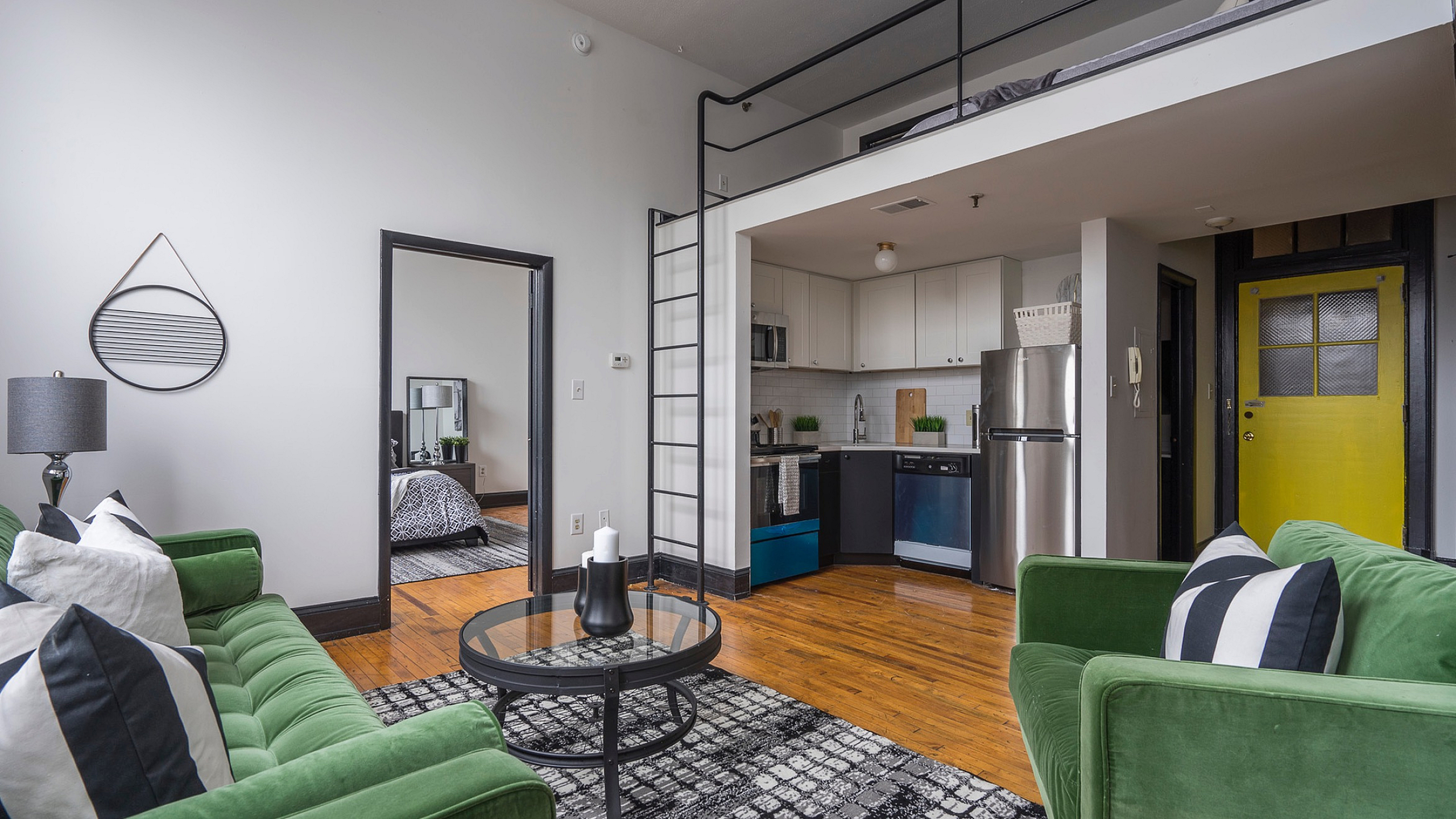 Chic loft apartment featuring a stylish living area with green velvet sofas and bold black-and-white accents, modern kitchen, and an inviting loft bed space. Ideal for contemporary urban living.