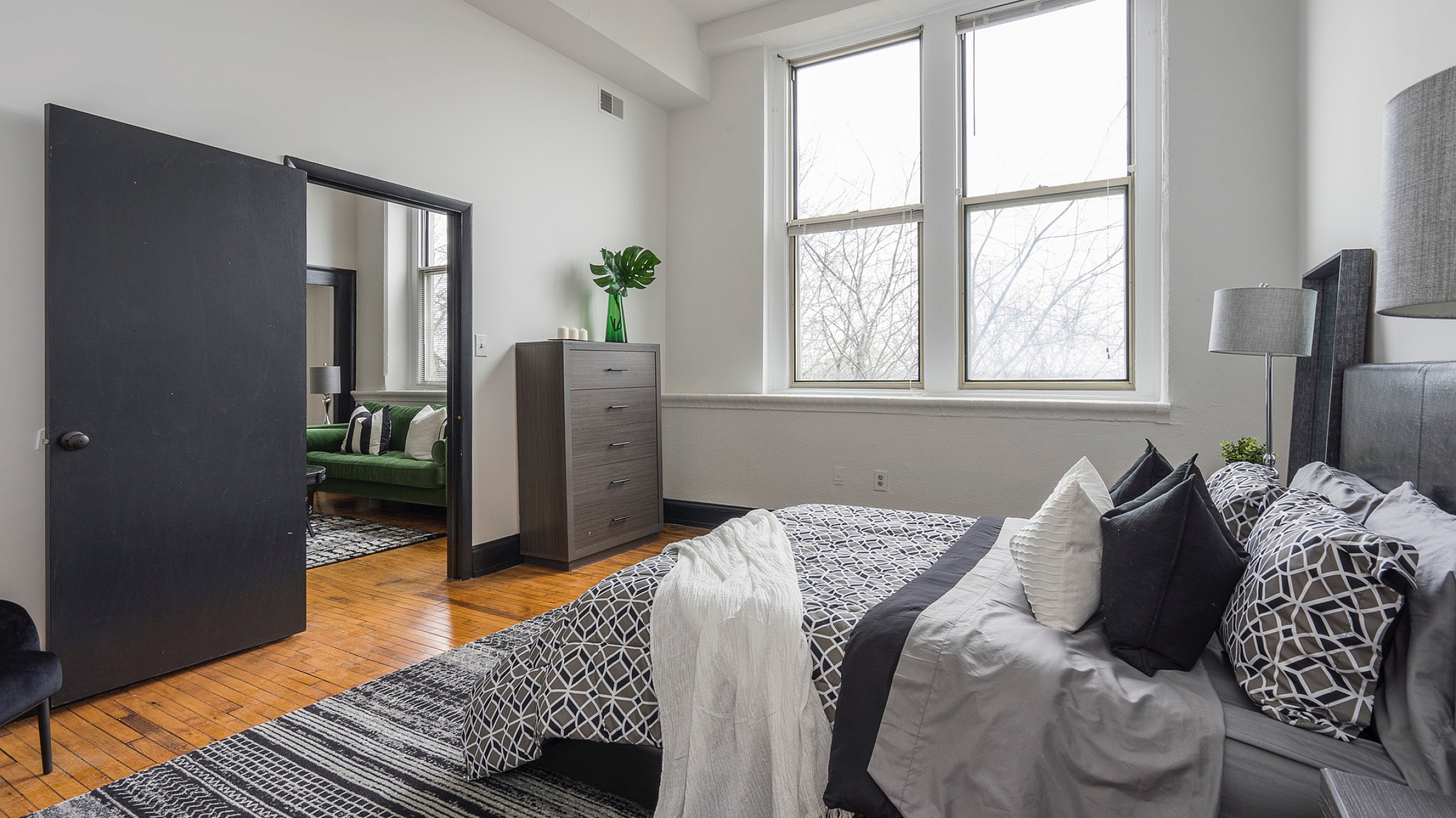 Bright and cozy bedroom with stylish decor, featuring a large window, elegant bed set, and sleek furniture, connecting to a modern sitting area.