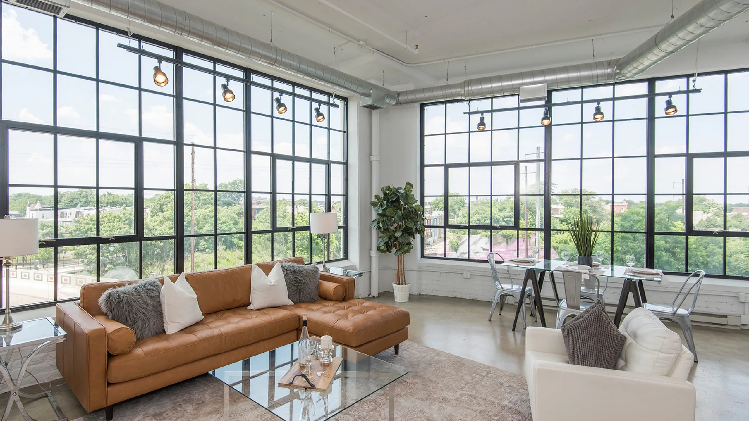 Bright and spacious loft with modern industrial design, featuring expansive windows, stylish seating, and a chic dining area with a lush green view.