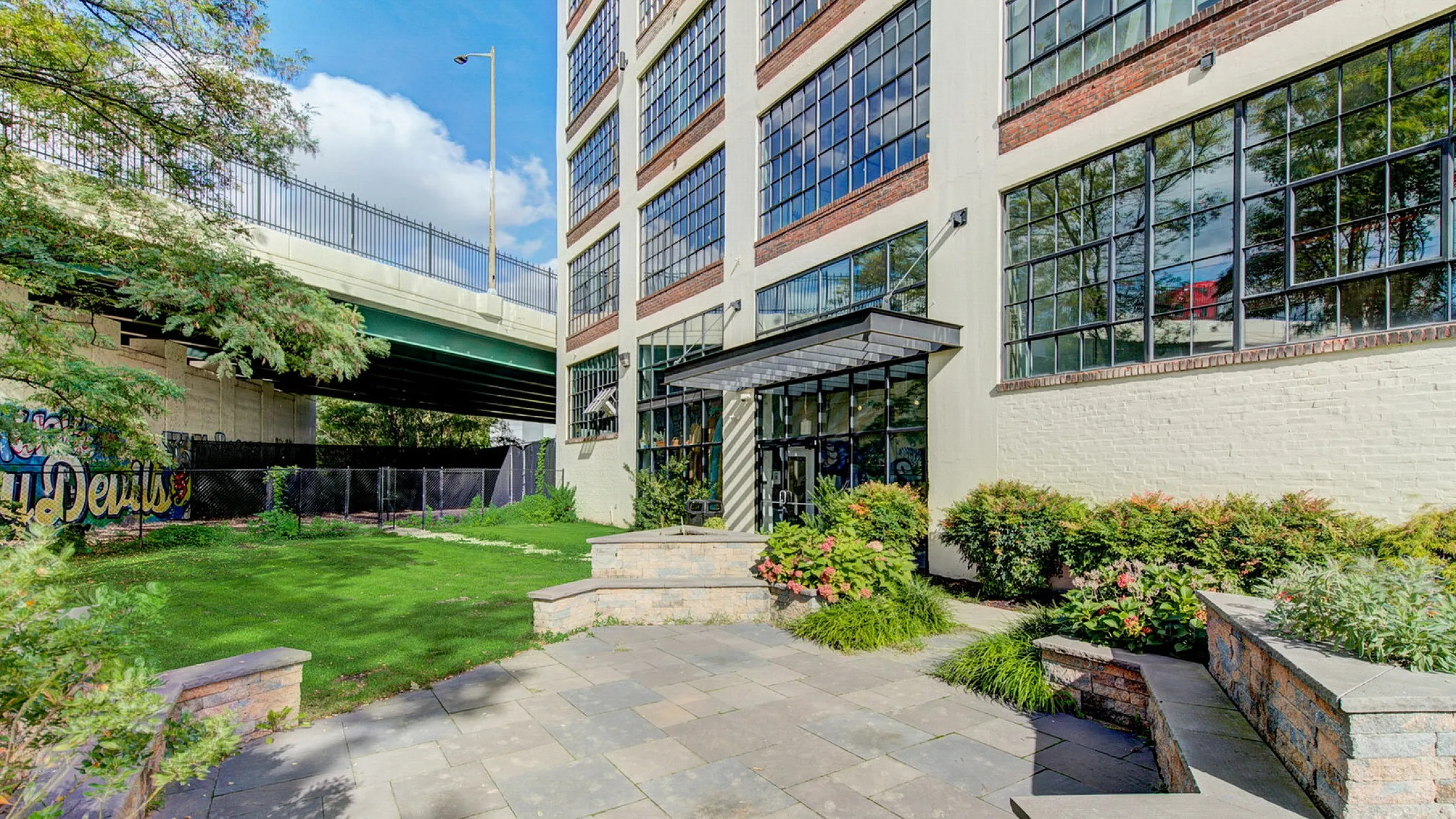 Modern apartment exterior with large industrial-style windows, lush greenery, and a spacious terrace under a clear blue sky.